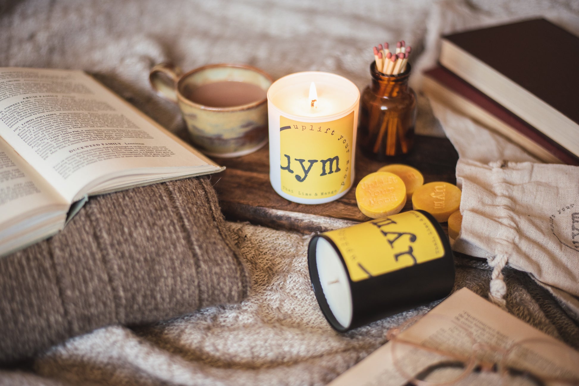 Thai Lime and Mango natural wax candle,matt black candle, matt white glass jar candle lighted, natural wax melts yellow in a cotton bag, books and a nice cup of tea, cosy atmosphere, Uplift Your Mood Scents